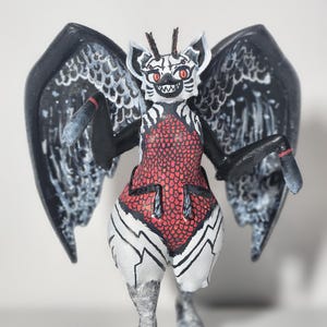 May include: A hand-painted figurine of a creature with large black wings, red eyes, and a white face with black markings. The torso features a red scale pattern, while the lower body is white with black lines. It has two small antennae.
