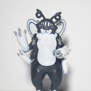 May include: A black and white creature figurine with four arms, large white fangs, multiple eyes, and insect-like wings. The figure has a textured surface, a white chest and limb accents, and stands on a circular base with a solid color background.