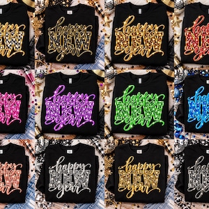 Op de afbeelding: Een collectie van twaalf zwarte sweatshirts, elk voorzien van de tekst "happy NEW year" in een uniek patroon of kleur, waaronder ruitjes, luipaardprint, rood, roze, paars, groen, blauw, roségoud, zilver en goudkleurige glitters.