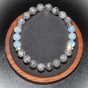 May include: A beaded bracelet featuring gray and white speckled stone beads, interspersed with light blue opal beads. Silver-toned spacers with clear crystals separate the bead types. The bracelet is displayed on a black surface within a wooden frame.