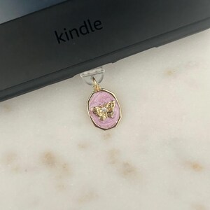 May include: A gold-colored butterfly charm with a pink background is attached to a black Kindle. The charm has a gold border and a small butterfly design with a clear gem in the center. The Kindle is black and has the word "kindle" printed on it.