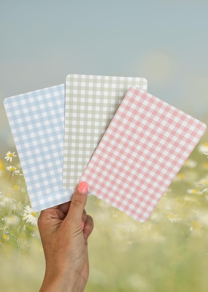 Puede incluir: Tres tarjetas rectangulares con un patr&oacute;n de cuadros vichy en colores pastel: azul claro, verde salvia y rosa. Las tarjetas se sostienen en una mano contra un fondo borroso de margaritas y un cielo azul suave.