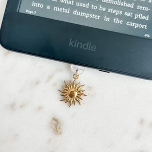 May include: A gold-colored sun charm attached to a Kindle e-reader. The e-reader displays text on its screen. The word "kindle" is printed on the device. The charm is hanging from the bottom of the e-reader.