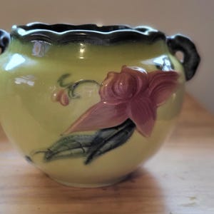 Peut inclure: Un petit pot en céramique vert avec deux anses. Le pot a un motif floral avec une fleur rose et des feuilles vertes.