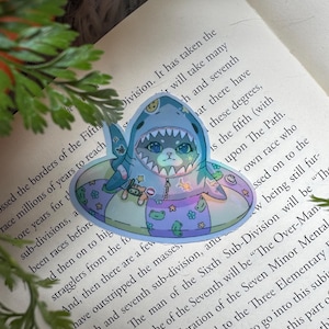 May include: A whimsical sticker featuring a cartoon animal wearing a shark costume and floating in a pool floatie. The sticker has a holographic effect and is decorated with stars, frogs, and other playful elements. The overall color scheme is pastel blue, purple, and green.