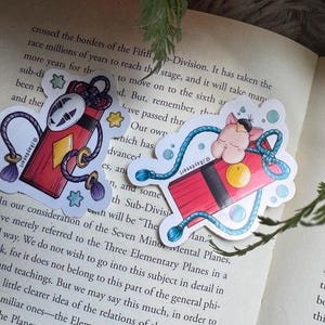 Puede incluir: Dos pegatinas caprichosas con un diseño rectangular rojo, un diamante, una máscara y un gato durmiente. Las pegatinas tienen acentos azules y morados, sobre una página de libro.