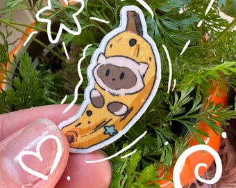 Puede incluir: Una pegatina con un personaje de dibujos animados disfrazado de plátano. El plátano es amarillo con manchas marrones y un tallo marrón. El personaje tiene una cara blanca y marrón y lleva un traje blanco. La pegatina se sostiene delante de una planta.