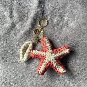 May include: A handmade crochet keychain featuring a starfish design in coral and white. The keychain includes a gold-colored ring and chain, with a small charm and a white seashell-shaped element. The background is a soft, gray fabric.