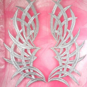 Pode incluir: Dois apliques de tecido decorativos de cor prateada em forma de asas estilizadas. Os apliques têm um design delicado e intrincado com uma aparência de renda.