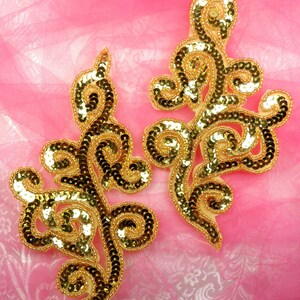 May include: Two gold sequined floral appliques on a pink fabric background. The appliques are made of small, round sequins sewn together in a floral pattern.