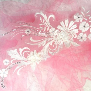 Peut inclure: Applique en dentelle blanche avec des paillettes et des embellissements nacrés sur un fond de tulle rose. L'applique présente des motifs floraux et des détails complexes.