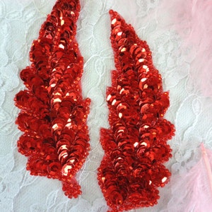 May include: Two red sequined feather shapes on a white lace background. The sequins are small and round, creating a shimmering effect.
