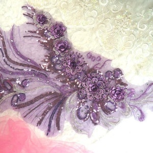 Peut inclure: Applique lavande avec des embellissements floraux et à paillettes. Le motif présente trois grandes fleurs détaillées et de plus petits accents floraux, avec des détails de paillettes argentées et violet foncé. L'applique est sur un tissu transparent violet clair.