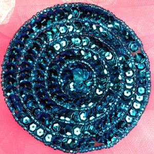 May include: A circular blue sequin embellishment with a concentric pattern. The sequins are arranged in a spiral design, creating a shimmering effect.