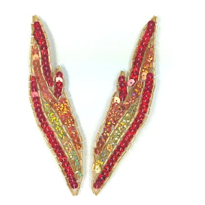 Może przedstawiać: Para czerwonych, pomarańczowych i złotych skrzydeł z cekinów. Skrzydła są wykonane z małych, opalizujących cekinów przyszytych do złotej tkaniny.