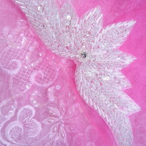 Puede incluir: Una aplicación de mariposa de cuentas blancas con un centro de strass, sobre un fondo de tela de tul rosa.