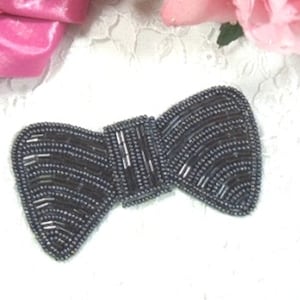 May include: A dark gray beaded bow tie. The bow is made of small, round beads that are sewn together to create a textured, three-dimensional effect.