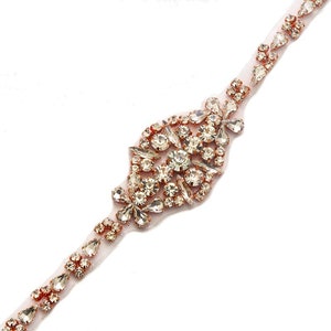 May include: A delicate rose gold beaded trim with a large, ornate rhinestone embellishment. The trim is perfect for adding a touch of sparkle to wedding dresses, bridal accessories, or other special occasion garments.