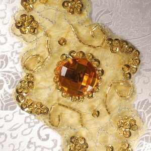 May include: A yellow beaded embellishment with a large, clear, orange rhinestone in the center. The embellishment is on a white fabric background.