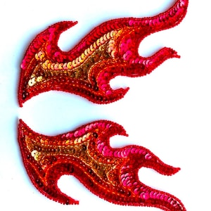 May include: Two red sequined flame patches. The flames are outlined in red and filled with red, pink, and gold sequins. The patches are designed to be sewn onto clothing or accessories.