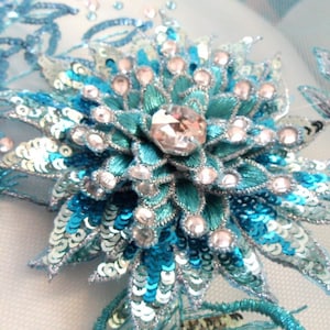 Pode incluir: Close-up de uma aplicação de flor de lantejoulas azuis e prateadas com um grande strass transparente no centro. A aplicação está sobre um fundo branco.