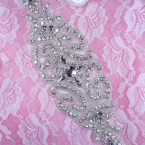 Peut inclure: Une embellissement en strass argenté avec un motif floral. L'embellissement est sur un fond de dentelle rose.