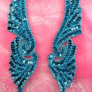 May include: Two blue sequined wings with a swirl design. The wings are on a pink background.
