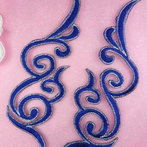 May include: Two blue embroidered appliques with silver trim. Each applique features a swirling, stylized design with curved lines and scroll-like elements. The appliques are set against a pink background.
