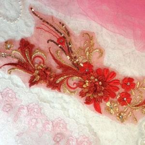Peut inclure: Applique florale rouge et dorée avec des paillettes et des strass. L'applique est sur un tissu rose transparent.