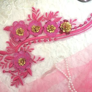 Peut inclure: Applique florale rose avec des accents dorés et des paillettes. L'applique est sur un fond blanc et présente un design incurvé avec plusieurs fleurs et feuilles.