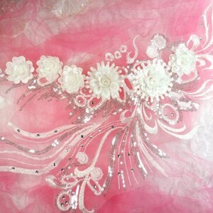 Peut inclure: Applique florale blanche avec des paillettes et des perles argentées sur un tissu de tulle rose. L'applique présente un groupe de fleurs blanches avec un motif floral délicat.