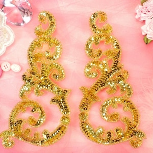 Könnte beinhalten: Zwei goldene Pailletten-Stoffapplikationen in Blumenform. Die Applikationen befinden sich auf einem rosa Hintergrund.