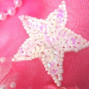 May include: A white star-shaped embellishment made of iridescent sequins and small white beads. The star is on a pink background.