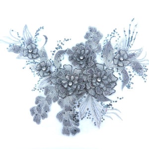Könnte beinhalten: Eine silberne Blumenapplikation mit Pailletten und Strasssteinen. Die Applikation zeigt eine Ansammlung von Blumen mit filigranen Details und einem zarten, femininen Design.