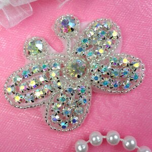 May include: A silver and iridescent butterfly-shaped embellishment with rhinestones and pearls. The embellishment is on a pink background.