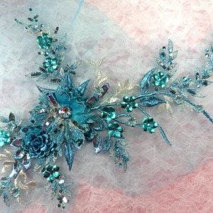 May include: Teal blue floral applique with sequins and beads. The applique features a large flower with smaller flowers and leaves. The applique is on a white background.