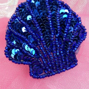 May include: A blue seashell-shaped applique made of sequins and beads. The applique is perfect for adding a touch of sparkle to crafts and clothing.