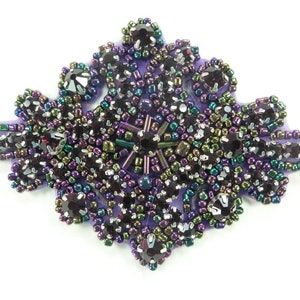 Può includere: Un abbellimento di perline viola, verde e nero con strass iridescenti. L'abbellimento ha la forma di un fiore con un'esplosione di stelle al centro.