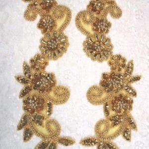 May include: A pair of symmetrical gold-colored embroidered appliqués featuring floral motifs, swirling vine patterns, and intricate beadwork. Each piece is crafted with metallic gold thread and shimmering sequins arranged in a dense, decorative design.