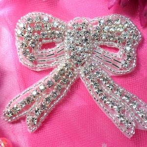 May include: A silver-colored bow with clear rhinestones and beads. The bow is on a pink background.