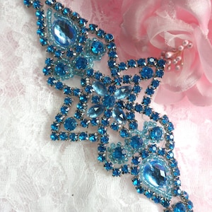 May include: A blue rhinestone embellishment with a teardrop shape and a starburst design. The embellishment is made of small, faceted rhinestones.