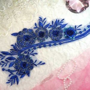 Peut inclure: Applique florale bleue avec des paillettes et des perles. L'applique est sur un fond blanc et présente un design incurvé avec cinq fleurs.