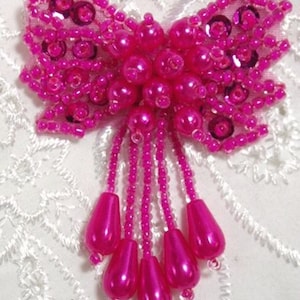 May include: A hot pink beaded bow brooch. The bow is made of small beads and sequins, with a cluster of larger beads at the centre. Dangling from the bow are beaded strands with teardrop-shaped beads. The brooch is set against a white lace background.