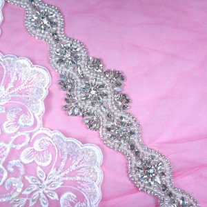 Peut inclure: Une garniture de perles argentées avec des strass clairs et des perles nacrées. La garniture est sur un fond de tissu rose.