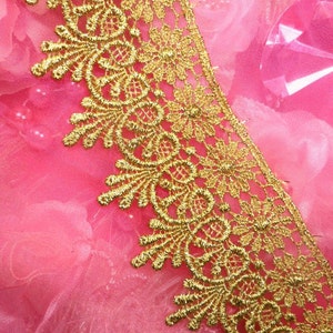 May include: Close-up of gold lace trim with intricate floral and scrollwork designs. The lace is set against a soft pink background, creating a contrast that highlights the detailed patterns. Ideal for sewing and craft projects.