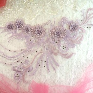 Peut inclure: Une applique florale lavande avec des paillettes et des strass. L'applique est sur un fond blanc.