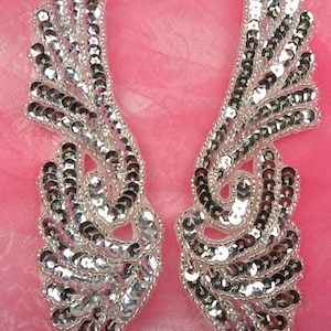 Puede incluir: Un par de alas de ángel de lentejuelas plateadas. Las alas están hechas de pequeñas lentejuelas redondas que están cosidas juntas en un patrón que se asemeja a plumas. Las alas están unidas a un respaldo de tela blanca.