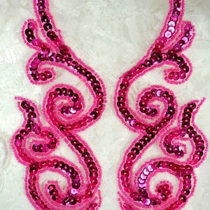 May include: Two pink sequined embellishments in a floral design. The embellishments are on a white lace background.