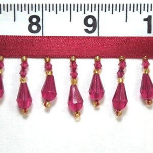 以下が含まれることがあります： 赤いビーズのトリムのクローズアップで、金色のアクセントが付けられています。トリムは、クリスタルのような外観の涙滴型のビーズの列を特徴としています。