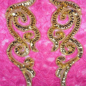 May include: Two gold sequined embellishments in a floral design on a pink fabric background.